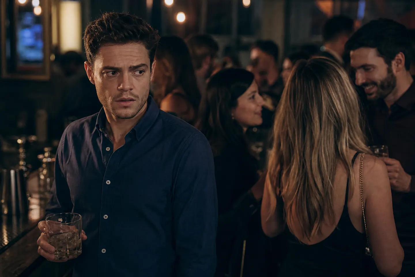 A man stands at the edge of a crowded bar holding a drink, looking tense while people chat in the blurred background