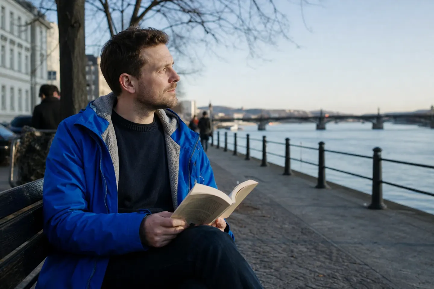 A man on a riverside bench in late afternoon looks up from a book toward the water, thinking