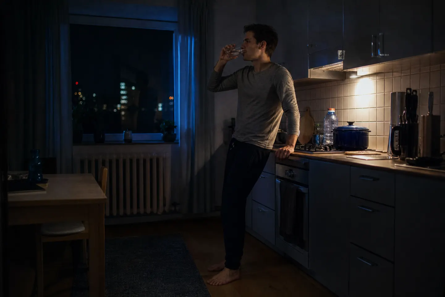 A man who cannot sleep drinks a glass of water in a dim kitchen late at night