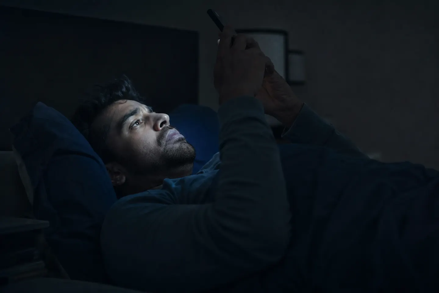 A man lies in bed at night scrolling his phone, the screen's glow on his face as he hesitates