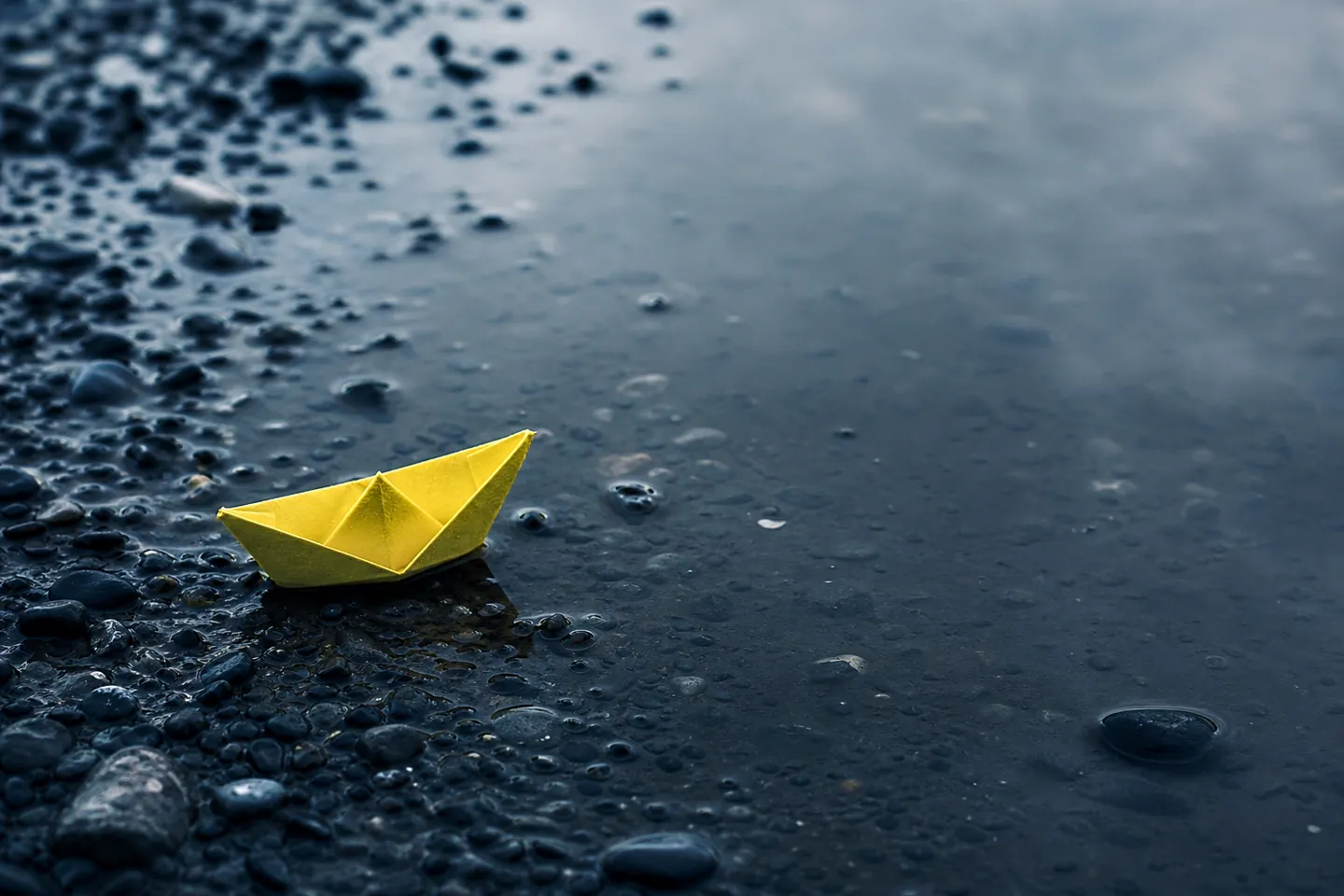 A yellow paper boat stranded on wet dark pebbles