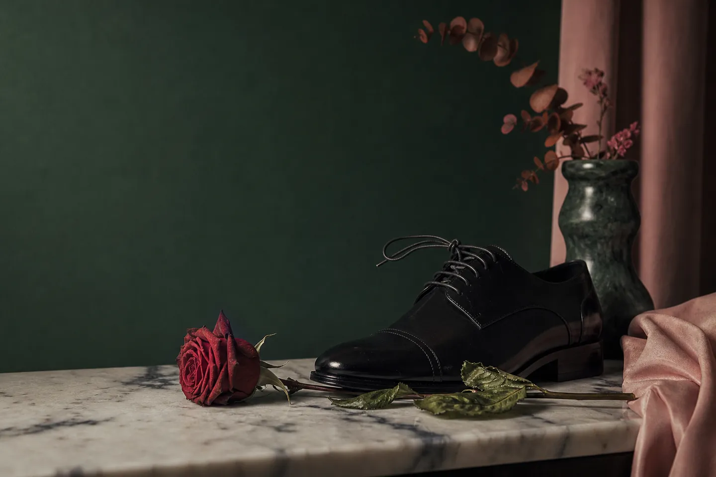 A polished dress shoe and a wilting red rose on marble against emerald green